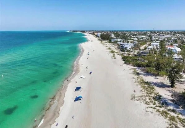 Vista aérea de una playa con aguas turquesas cristalinas, una orilla arenosa y una zona residencial con casas y vegetación.