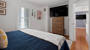 A bedroom with a bed, a wooden dresser, a television mounted on the wall, and a window with blinds.