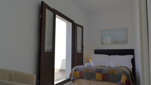 Ein Schlafzimmer mit Bett, einem Sessel, einem Bild an der Wand und einer Tür, die zu einem Außenbereich führt.