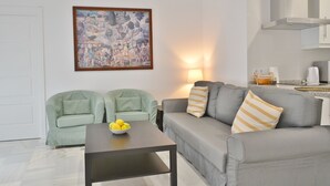 Una sala moderna con un sofá gris, dos sillones verdes, una mesa de centro con limones, un cuadro en la pared y una zona de cocina al fondo.