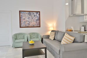 Una sala moderna con un sofá gris, dos sillones verdes, una mesa de centro con limones, un cuadro en la pared y una zona de cocina al fondo.