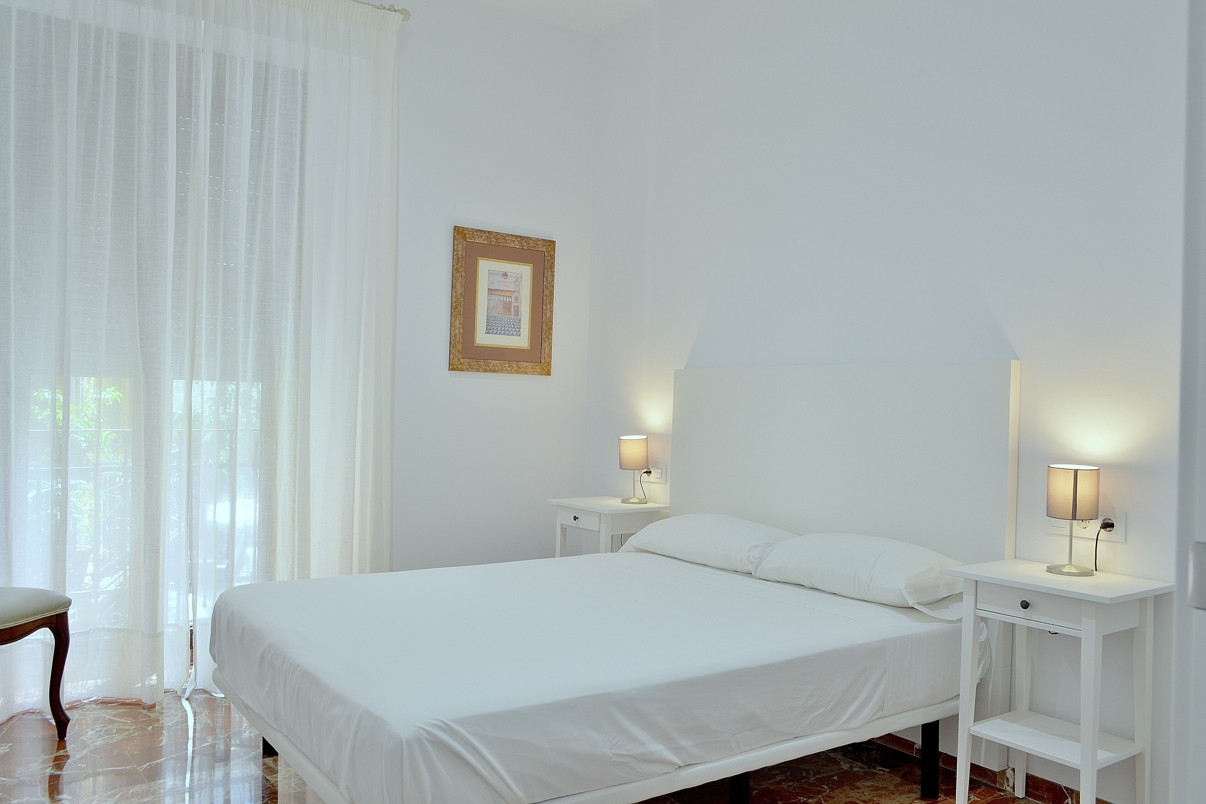 Un dormitorio con una cama, dos mesitas de noche, una silla y un cuadro en la pared.
