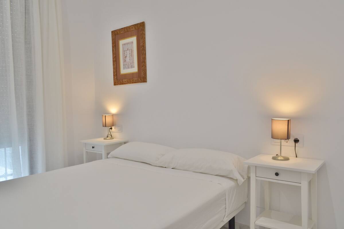 Una cama bien hecha con sábanas blancas, dos mesitas de noche con lámparas, un cuadro enmarcado en la pared y una ventana con cortinas de encaje.
