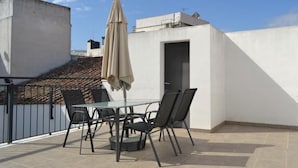 Una terraza en la azotea con una mesa y sillas de vidrio, un paraguas color beige y una pared blanca.