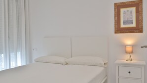 Un dormitorio con cama, cortinas blancas, lámpara de noche, un cuadro en la pared y una mesita de noche blanca con cajones.