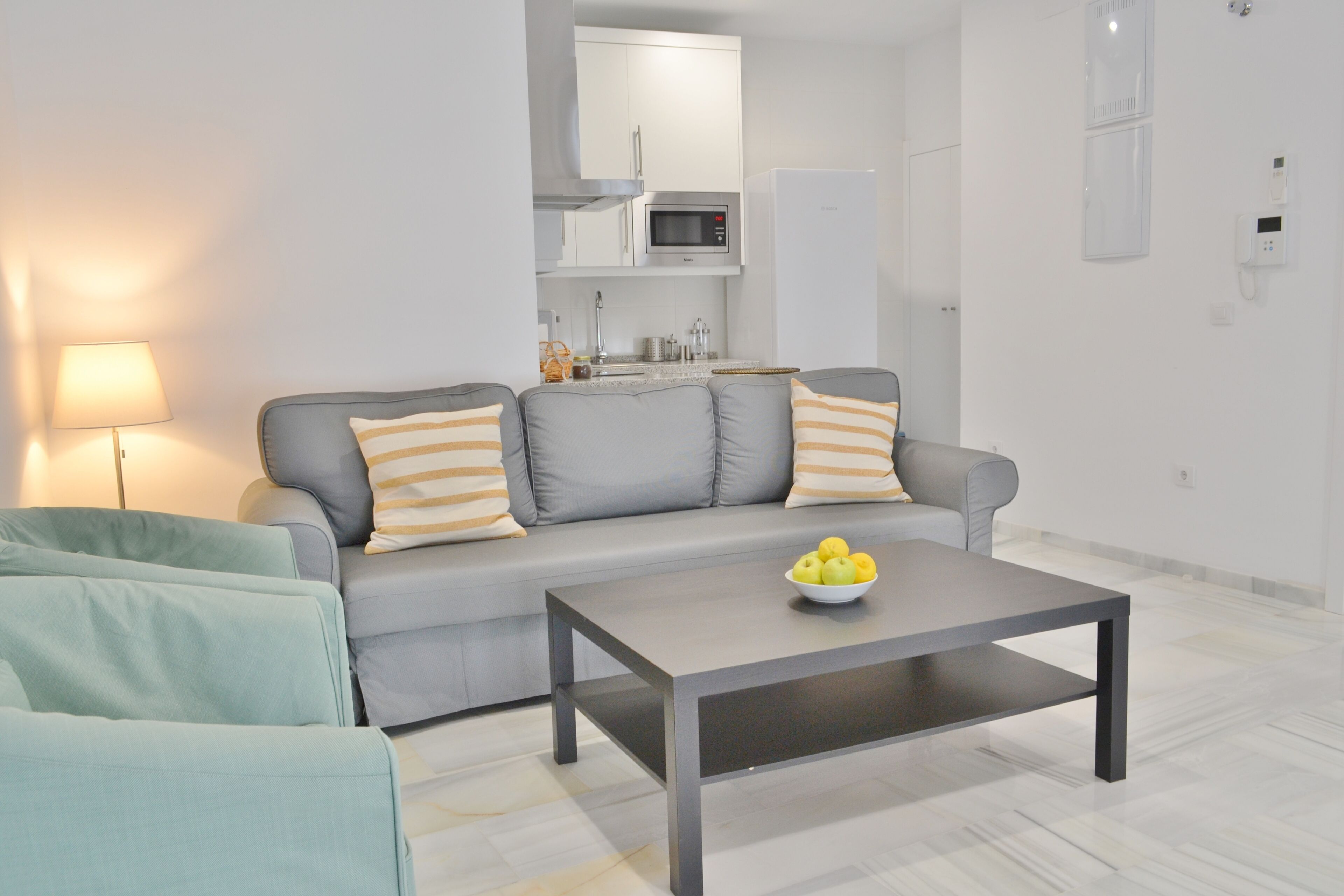 Una sala de estar moderna con un sofá gris, una mesita de centro y una cocina al fondo.