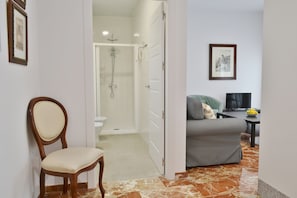 Una habitación con ducha, una silla, un sofá, una mesa con un frutero y un cuadro en la pared.