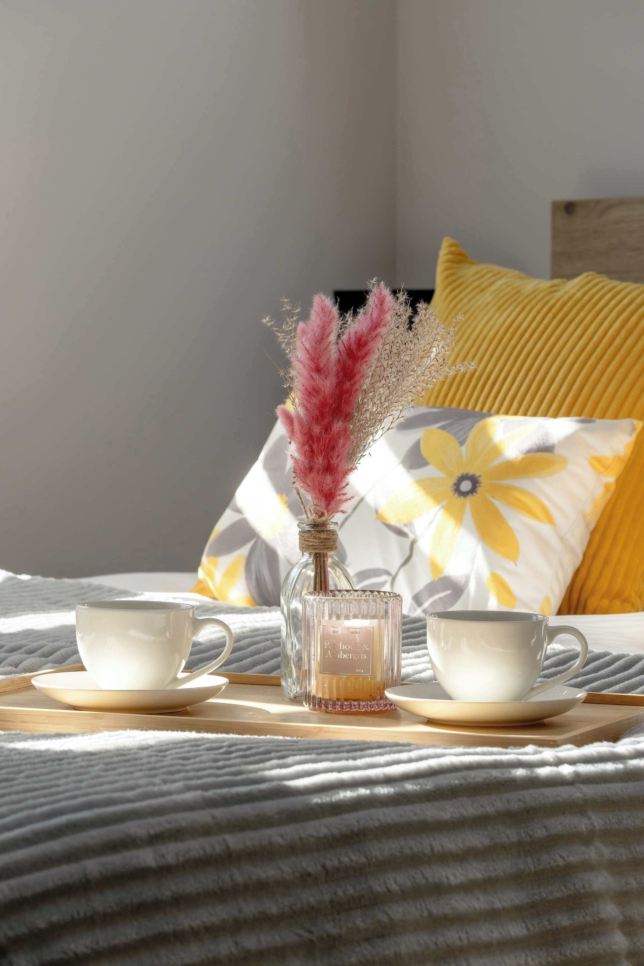 A bed with two white cups and saucers, a clear glass bottle with pink feathers, and a yellow pillow with a floral design.