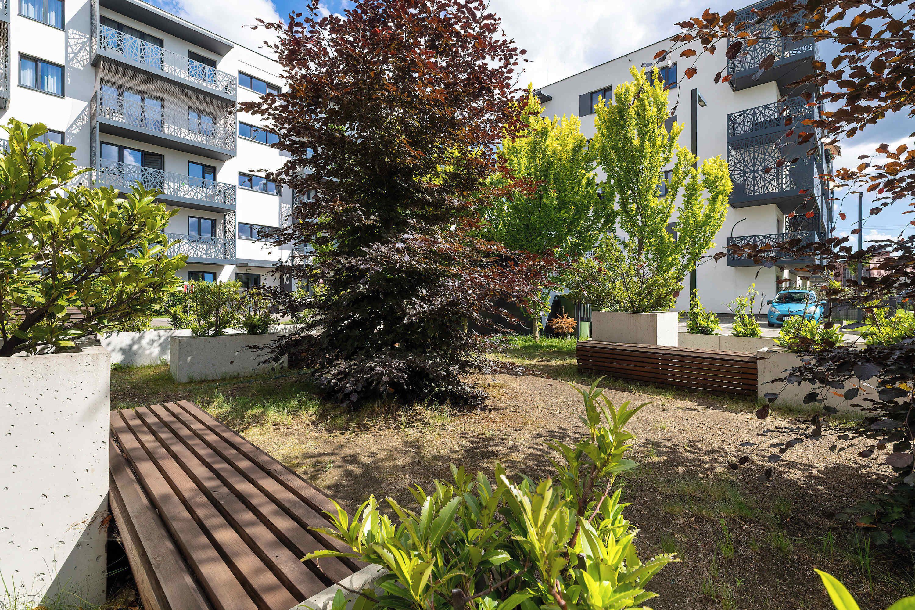 A modern apartment complex with balconies, a wooden bench, and a variety of trees.