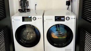 A laundry room with two front-loading washing machines, a shelf with cleaning supplies, and a power outlet. - Brick & Bed - Riverfront Loft (St. Louis)