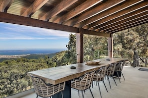 An outdoor dining area with a long wooden table and wicker chairs, overlooking a scenic landscape.