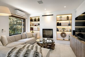 A modern living room with a sofa, coffee table, and fireplace. There are shelves with decorative items and a TV on a wooden cabinet.