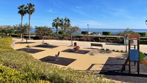 Un parque con bancos, juegos infantiles, palmeras y vistas al mar.