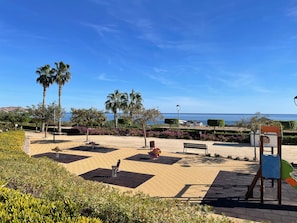 Ein Park mit Bänken, Spielgeräten, Palmen und Blick auf das Meer.