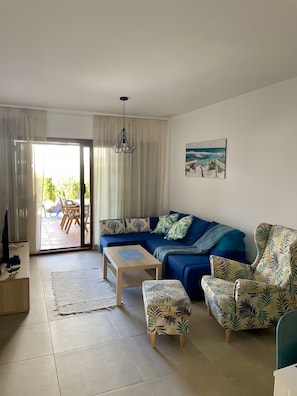Ein Wohnzimmer mit blauem Sofa, einem gemusterten Sessel, einem Couchtisch und Blick ins Freie durch eine Glastür.