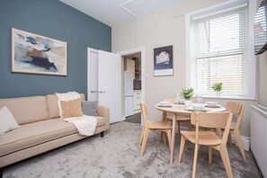 A modern living room with a beige sofa, a dining table with chairs, a kitchen in the background, and a large abstract painting on the wall.
