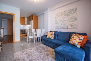 Una sala de estar moderna con un sofá azul, una zona de comedor con sillas blancas y una cocina al fondo.