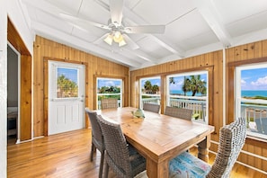 A dining area with a wooden table and chairs, a ceiling fan, and large windows with views of the ocean. - Welcome to Sisters By The Sea! (St. George Island)
