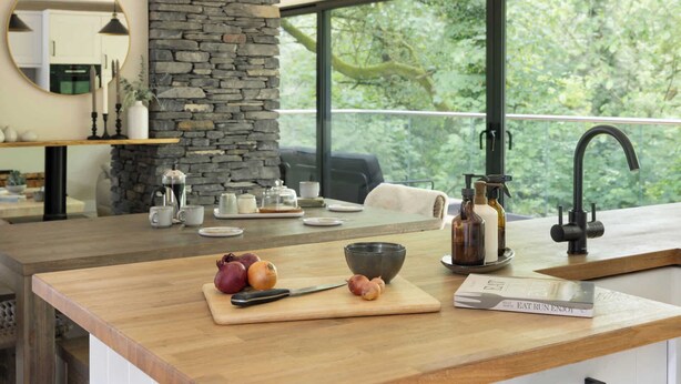 A modern kitchen with a stone wall, large windows, and a wooden countertop with a cutting board, knife, and book. - Fawns Keep - Hot Tub & Sauna Retreat | Ridgestone (Bowness-on-Windermere)