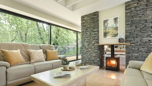 A modern living room with a stone fireplace, a sofa, a coffee table, and a view of greenery outside. - Fawns Keep - Hot Tub & Sauna Retreat | Ridgestone (Bowness-on-Windermere)