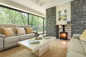 A modern living room with a stone fireplace, a sofa, a coffee table, and a view of greenery outside. - Fawns Keep - Hot Tub & Sauna Retreat | Ridgestone (Bowness-on-Windermere)