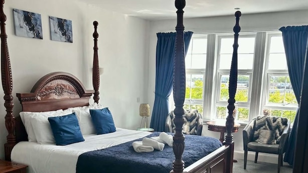 A four-poster bed with a headboard, a bedside table, a chair, and a view of the outdoors through large windows.
