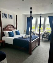 A four-poster bed with a headboard, a bedside table, a chair, and a view of the outdoors through large windows.