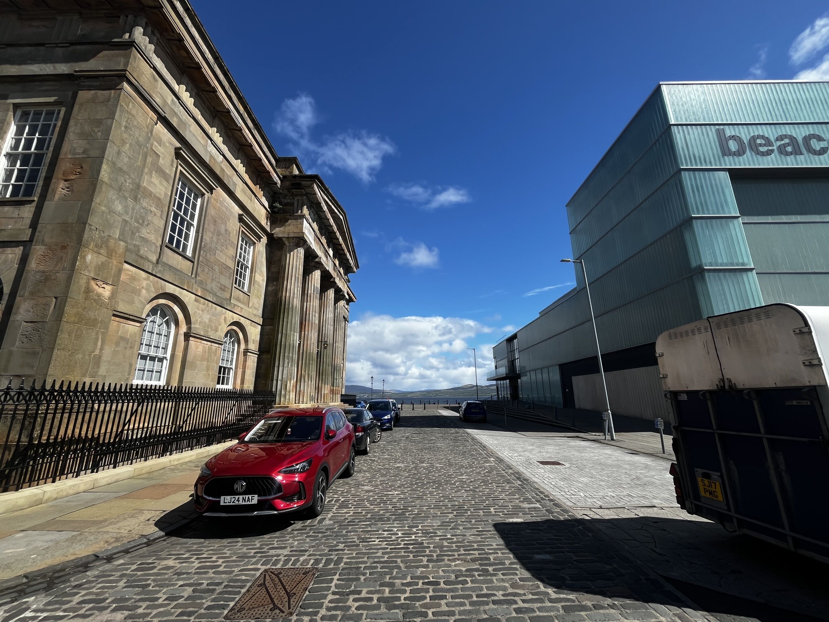 Une rue pavée avec une voiture rouge garée sur le bord, un bâtiment historique orné de colonnes et un bâtiment moderne dont la façade porte l'inscription « beac ».