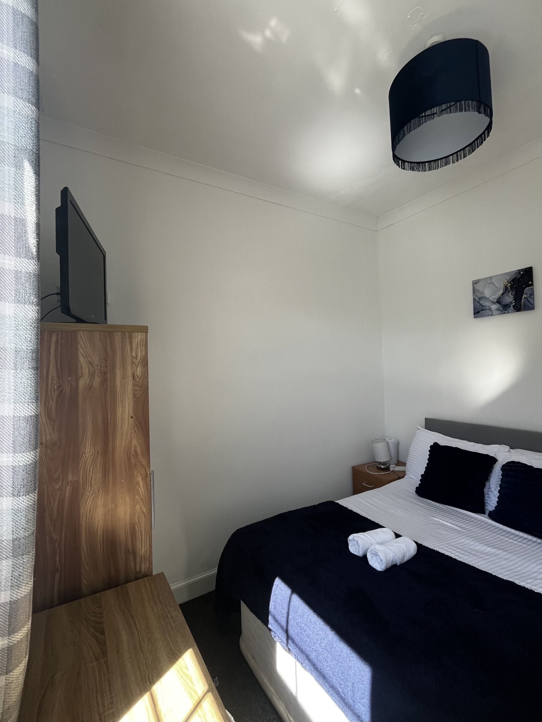 Un dormitorio con cama, mesita de noche, lámpara, un cuadro en la pared y ventana.
