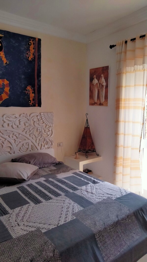 Une chambre à coucher avec un lit, des oreillers, une tête de lit décorative, une décoration murale, une petite table avec une bougie et une fenêtre avec des rideaux.