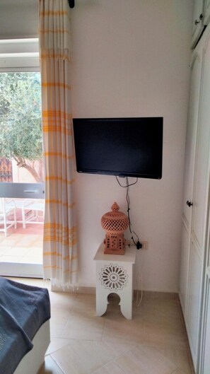 Une pièce avec un téléviseur fixé au mur, une petite table blanche agrémentée d’un objet décoratif, et une vue sur l’extérieur par une fenêtre ornée de rideaux.