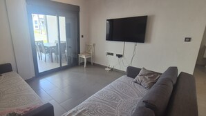 Ein Wohnzimmer mit zwei Sofas, einem an der Wand montierten Fernseher und einer Glastür, die zu einem Außenbereich mit Sitzgelegenheiten führt.