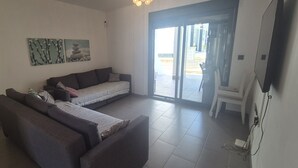Ein Wohnzimmer mit einer Couch, einem kleinen Tisch und einem Fernseher. Eine Glasschiebetür führt nach draußen.