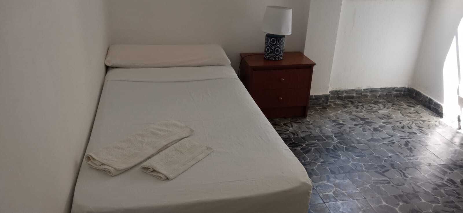 Un lit simple avec un matelas blanc et deux serviettes pliées, une table de chevet avec une lampe et un sol carrelé en pierre.