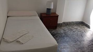 Un lit simple avec un matelas blanc et deux serviettes pliées, une table de chevet avec une lampe et un sol carrelé en pierre.