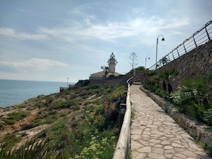 Un paseo marítimo que conduce a un faro, con un muro de piedra a un lado y una barandilla metálica al otro.