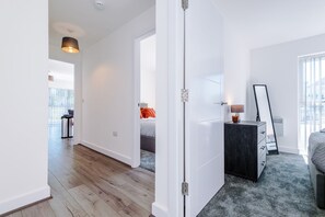 Ein modernes Hotelzimmer mit Holzboden, einem Bett mit grauer Bettdecke, einem Nachttisch mit Lampe und einem großen Fenster mit Jalousien.