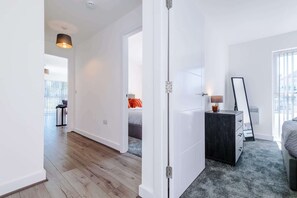 A modern hotel room with a wooden floor, a bed with grey bedding, a dark dresser, and a large window with blinds. - Stylish 2BDR Hospitals Nearby Free Parking (Liverpool)