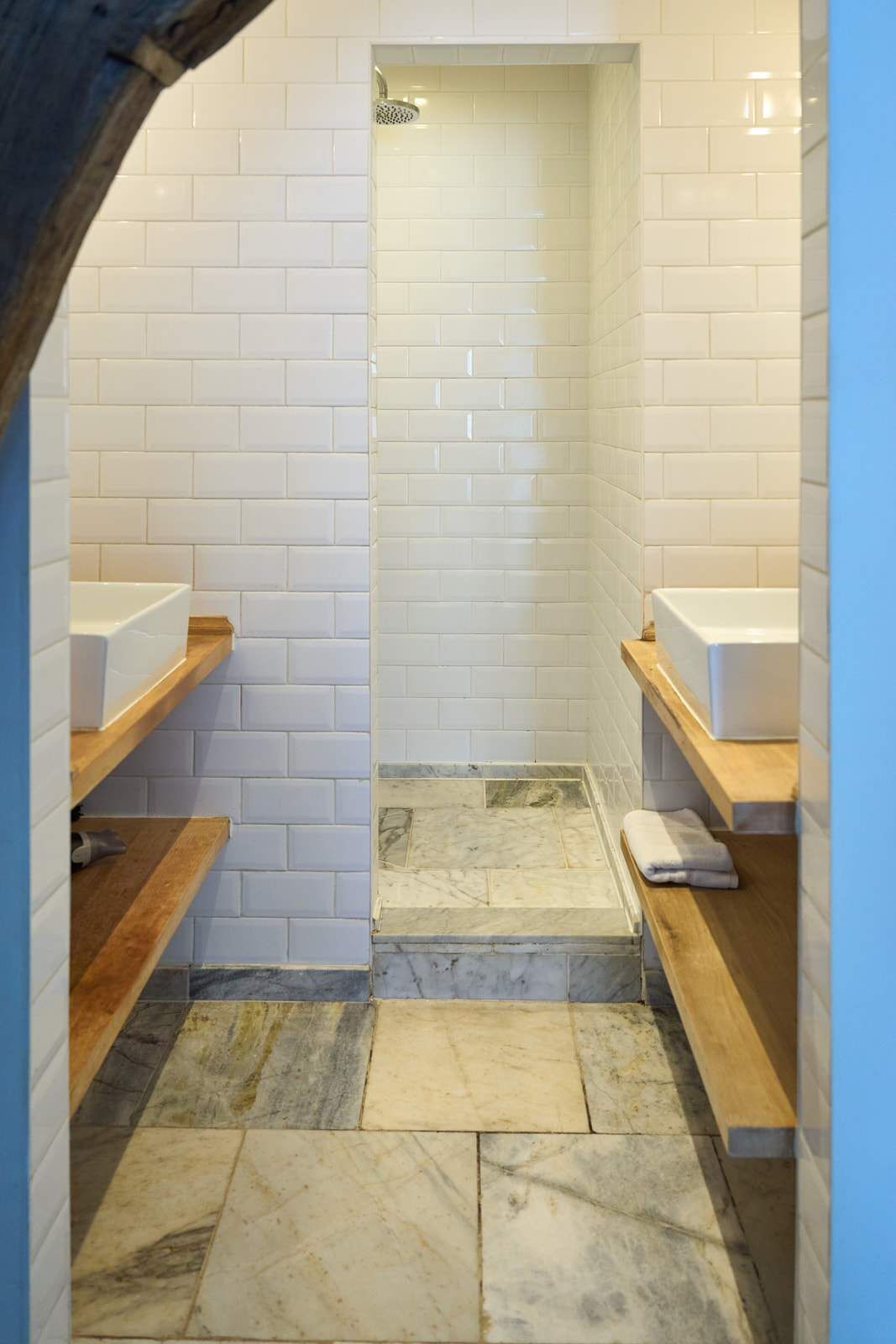 Une salle de bain avec des murs recouverts de carreaux blancs, un sol en marbre et des étagères en bois.