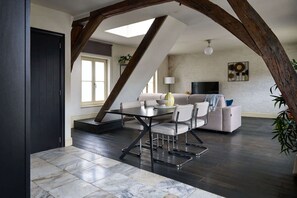Ein Loft-Zimmer mit hohen Decken, freiliegenden Holzbalken, einem Esstisch mit Stühlen, einer Couch, einem Fernseher und einer Pflanze.