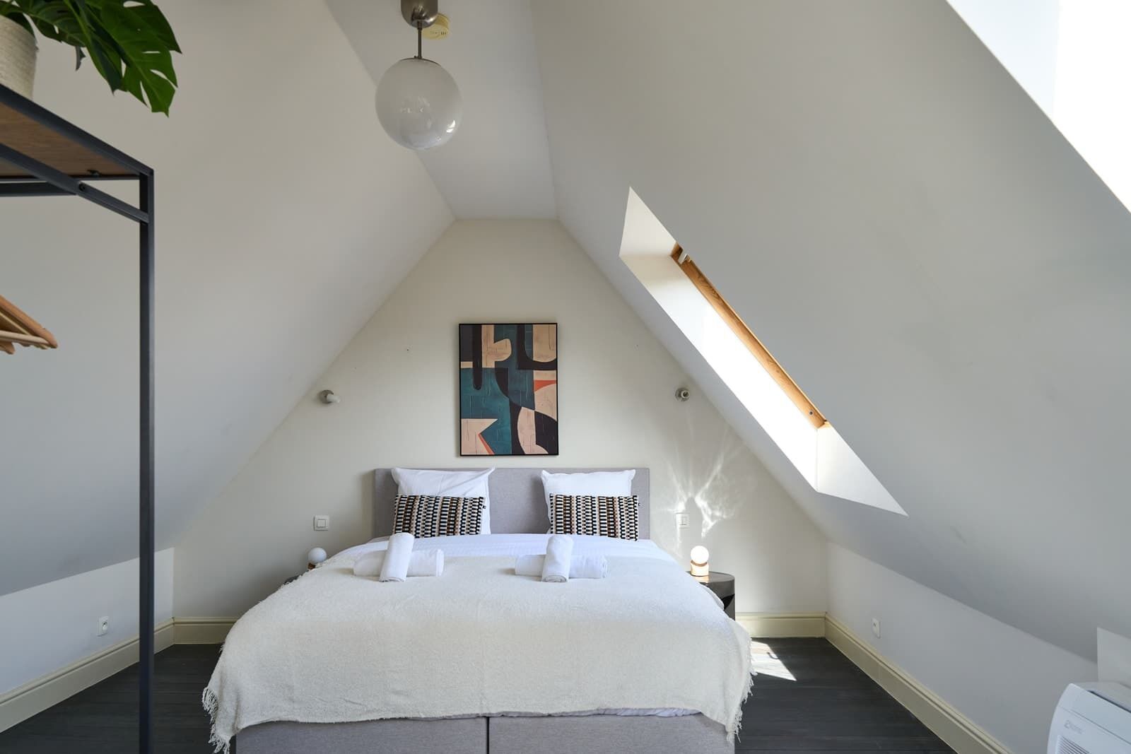 Ein Schlafzimmer mit schrägen Wänden, einem Bett mit weißer Bettwäsche, einem geometrischen Wandbild und einem Dachfenster.