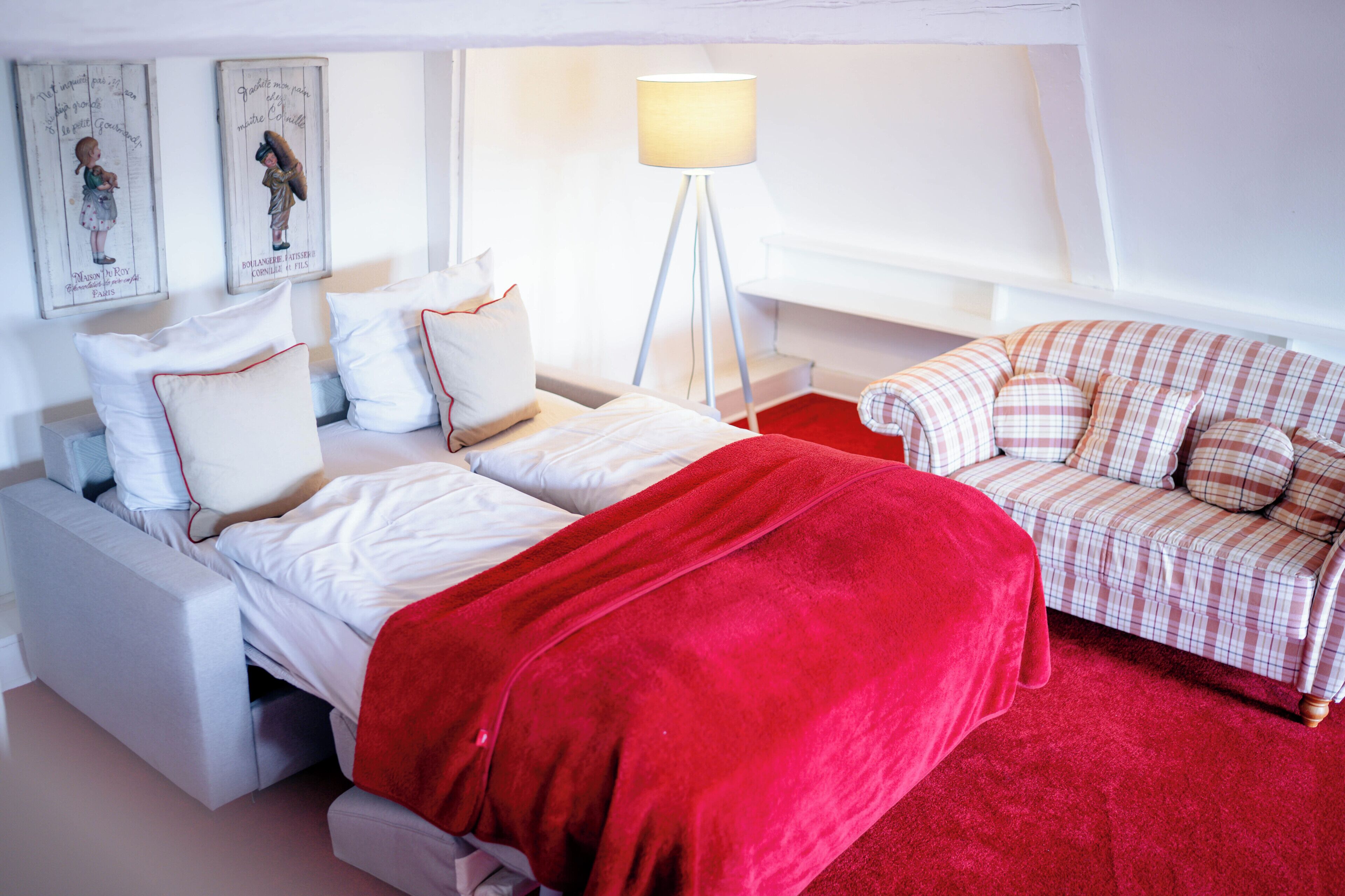 A bedroom with a bed, a red blanket, a white sofa, and a floor lamp.