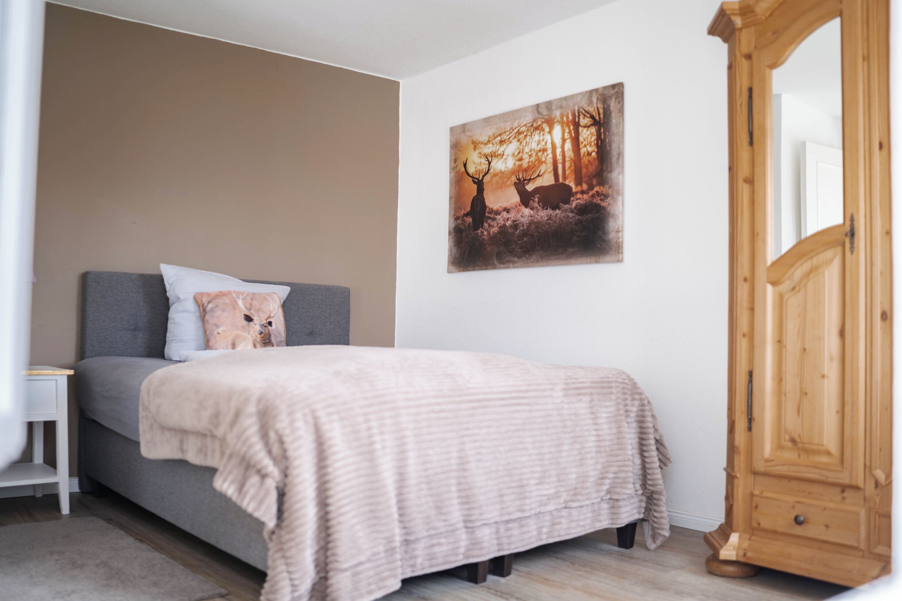 A bedroom with a bed, a nightstand, a wooden wardrobe, and a deer painting on the wall.