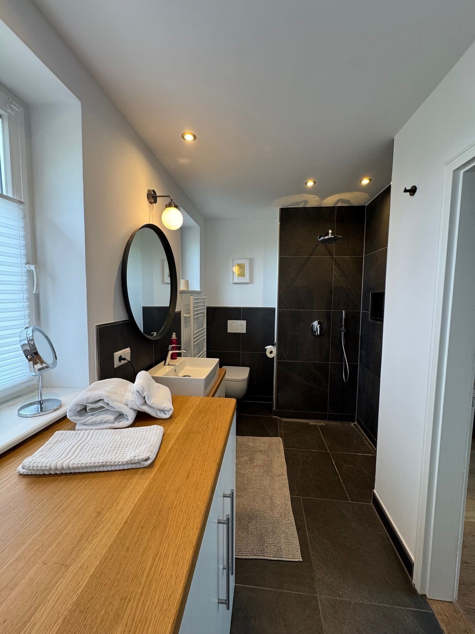 Ein modernes Badezimmer mit einem Holzkosmetikschrank, einem runden Spiegel, einer Toilette und einem dunklen Duschbereich.
