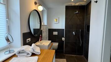 Ein modernes Badezimmer mit einem Holzkosmetikschrank, einem runden Spiegel, einer Toilette und einem dunklen Duschbereich.