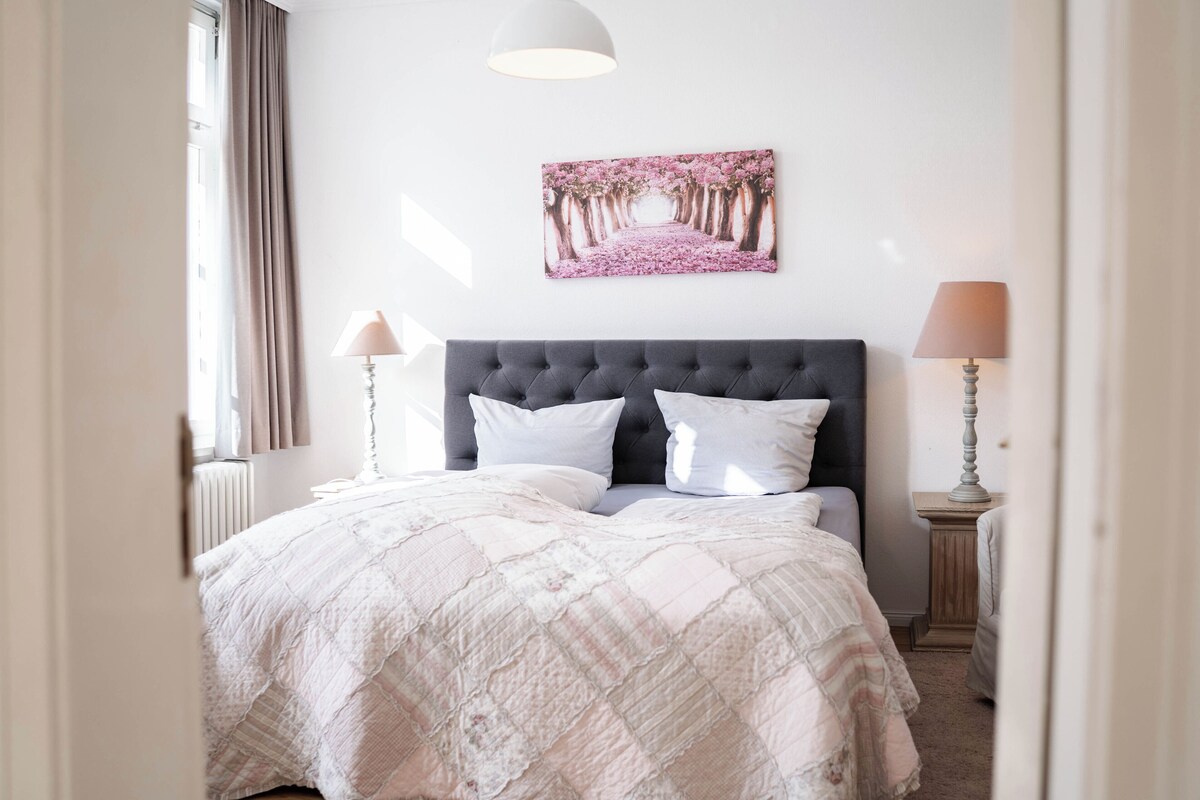 Un dormitorio con una cama grande, un cabecero, dos lámparas de noche, una ventana con cortinas y un cuadro colgado en la pared.