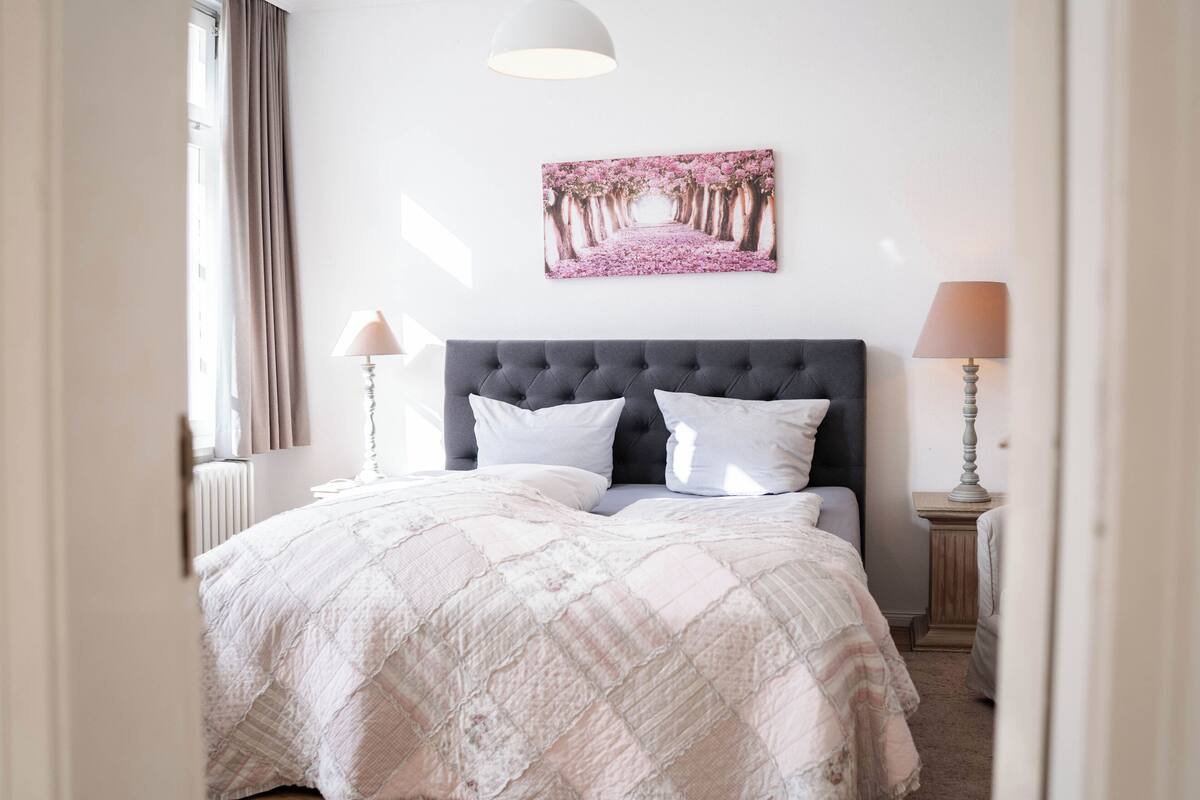 Ein Schlafzimmer mit einem großen Bett, einem Kopfteil, zwei Nachttischlampen, einem Fenster mit Vorhängen und einem an der Wand hängenden Bild.