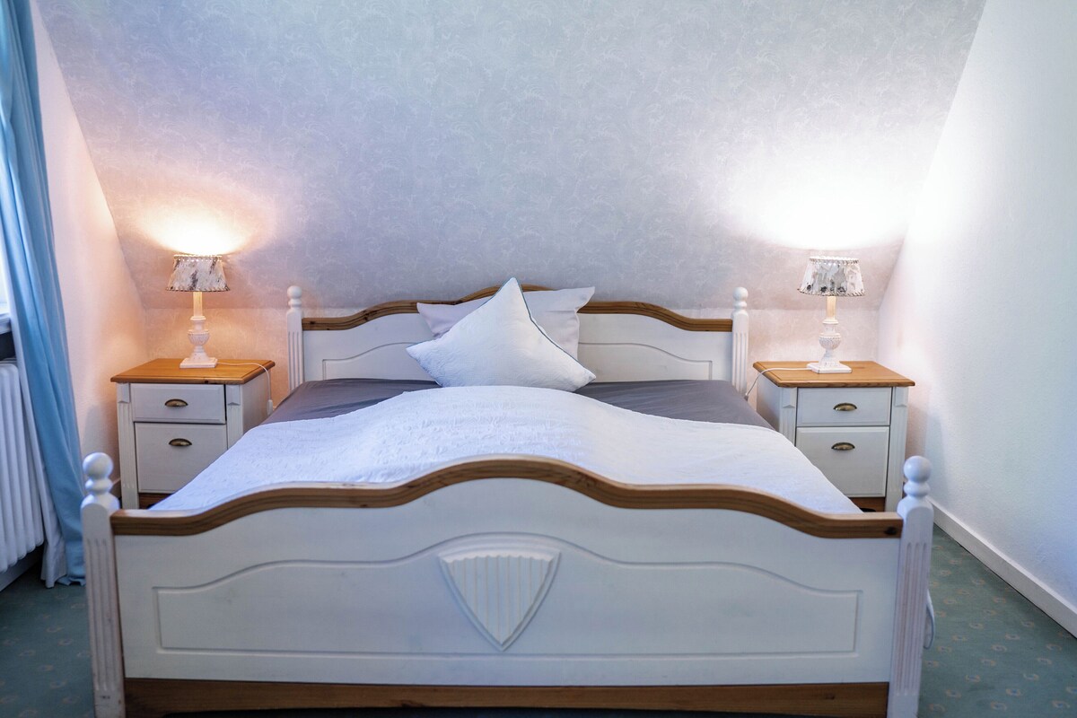 Una cama bien hecha con ropa blanca, dos mesitas de noche con lámparas y un papel tapiz estampado.