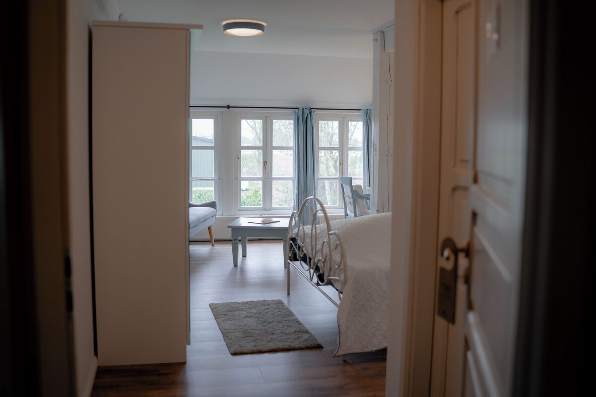 Una habitación con cama, mesita de noche, ventana con cortinas y una puerta.
