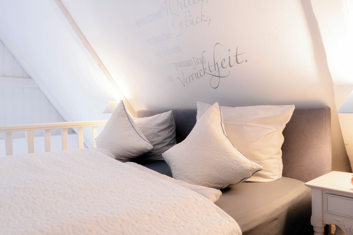 Un dormitorio con techo inclinado, una cama con ropa blanca y una mesita de noche con lámpara.